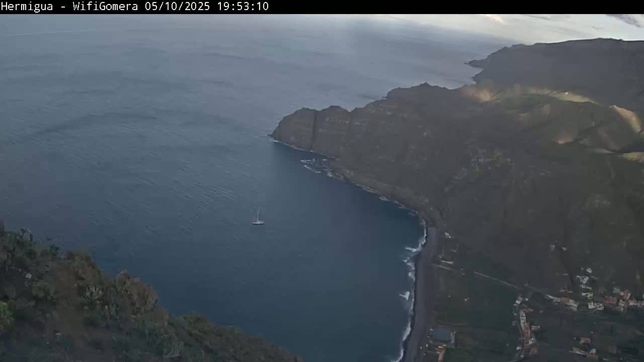 A sailboat is visible in a calm, dark blue ocean bay nestled between dark, rocky cliffs under a mostly clear sky.
