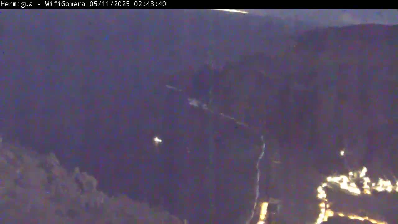 A dark, nighttime view of a hilly landscape with a few scattered lights visible in the distance.