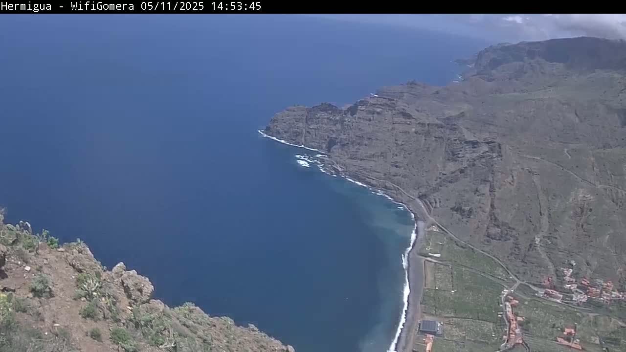 A high-angle view shows a dark-blue ocean meeting a rocky, dark-grey coastline under a partly cloudy sky.