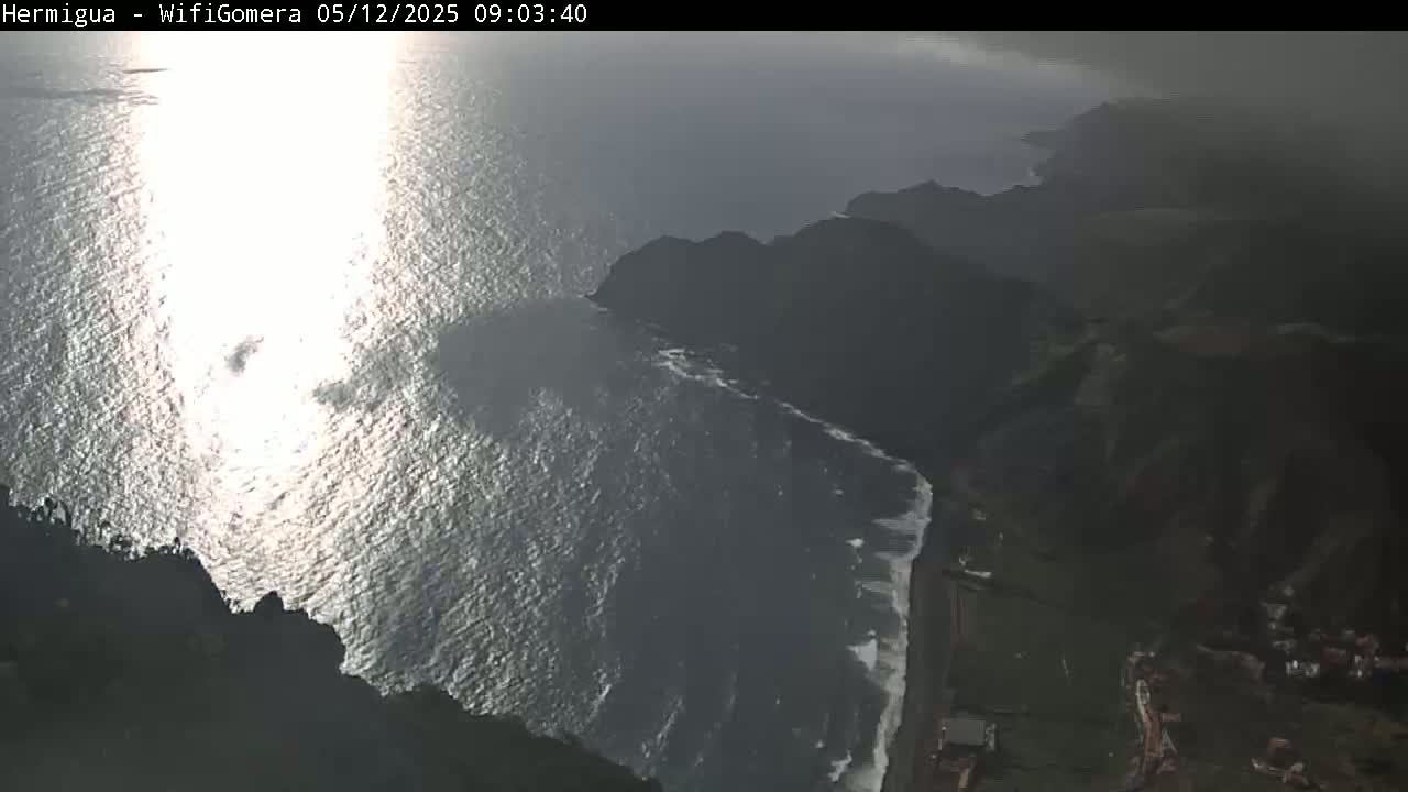 A sunlit ocean meets a dark, rocky coastline with a small town visible in the lower right corner, under somewhat overcast skies.