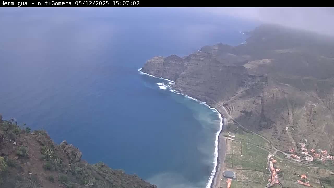 A hazy, high-angle view shows dark, rocky cliffs meeting a dark-blue ocean with a small, sandy beach and some low-lying buildings.