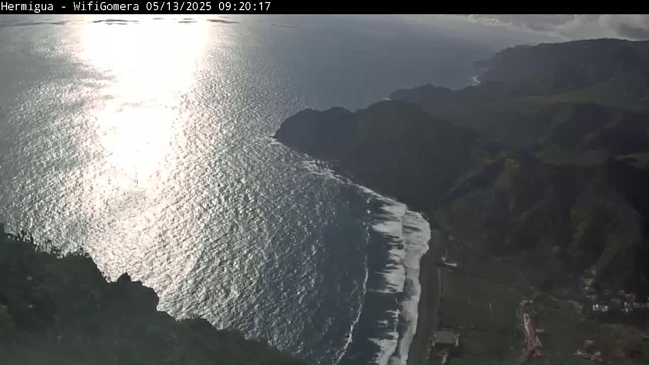 A bright, sunny day shows a rocky coastline with dark volcanic hills meeting a sparkling ocean, where small waves break on a dark sand beach.