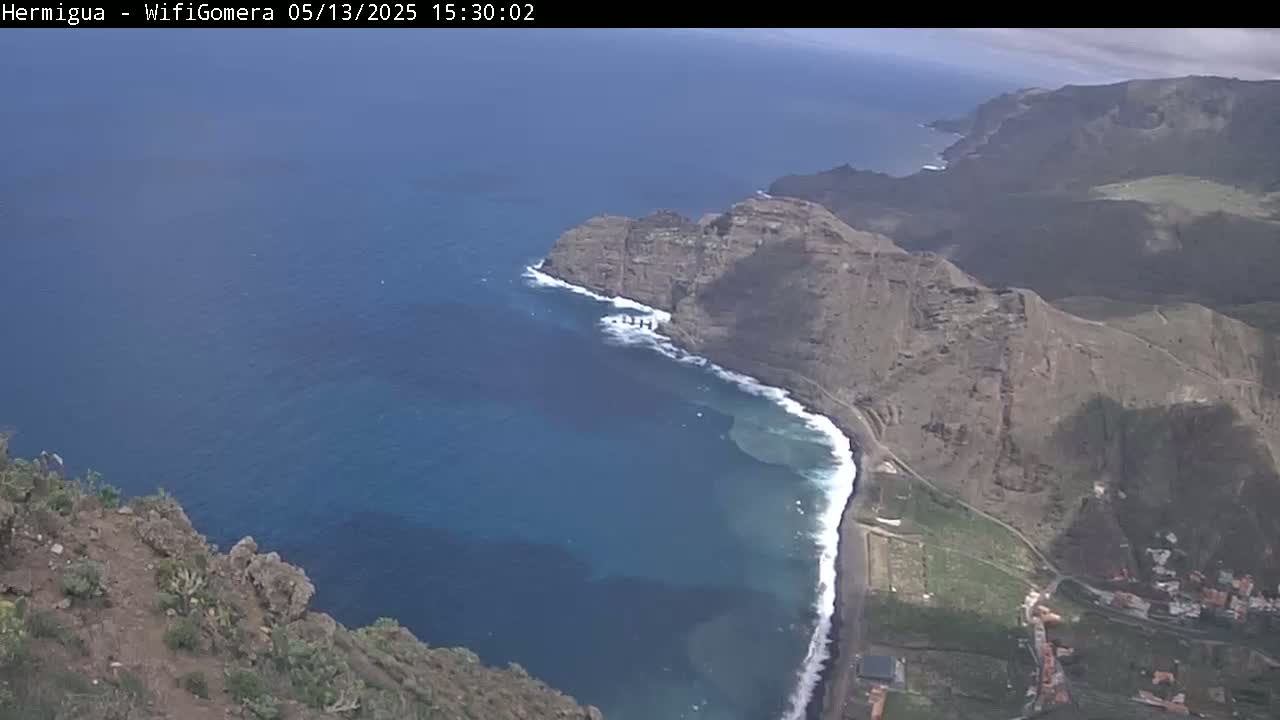 A high-angle view shows a dark-blue ocean meeting a rocky, dark-brown coastline with a small amount of green vegetation under mostly sunny skies.