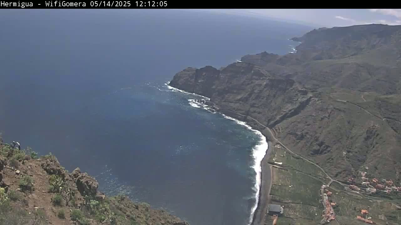 A high-angle view shows a rocky coastline meeting a dark-blue ocean under a bright, sunny sky.
