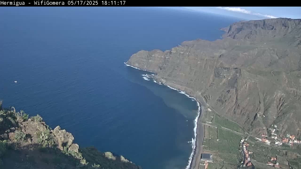 A high-angle view shows a rocky, mountainous coastline meeting a dark blue ocean on a sunny day; a small village is visible near the water's edge.