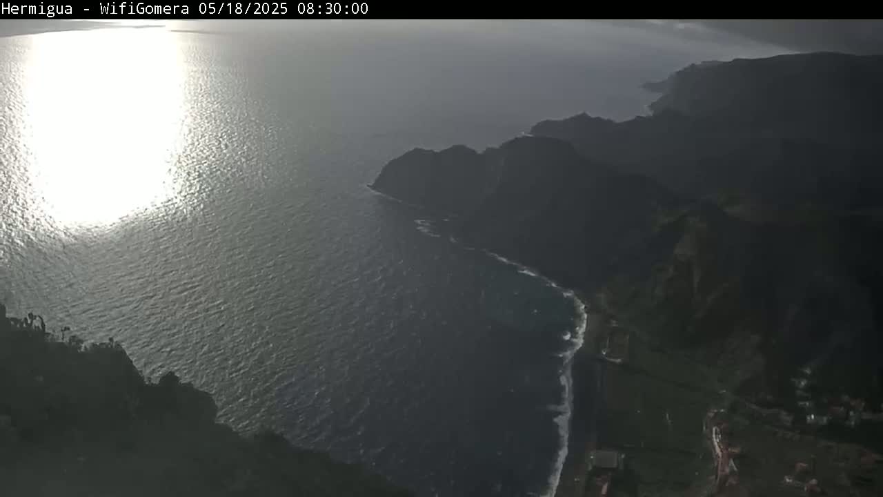A high-angle view shows a dark, rocky coastline meeting a sunlit ocean under a mostly cloudy sky.