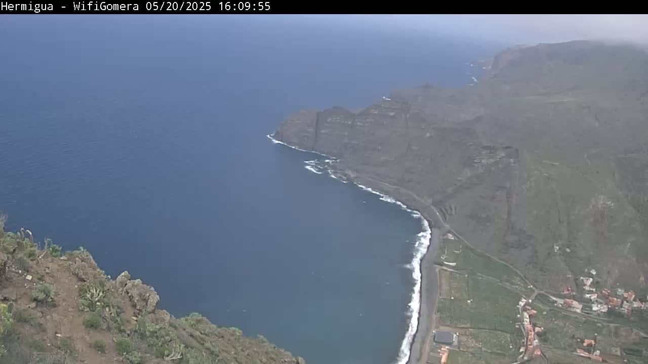 A high-angle view shows a dark-blue ocean meeting a rocky, dark-grey coastline with a small, sandy beach, under hazy or overcast conditions.