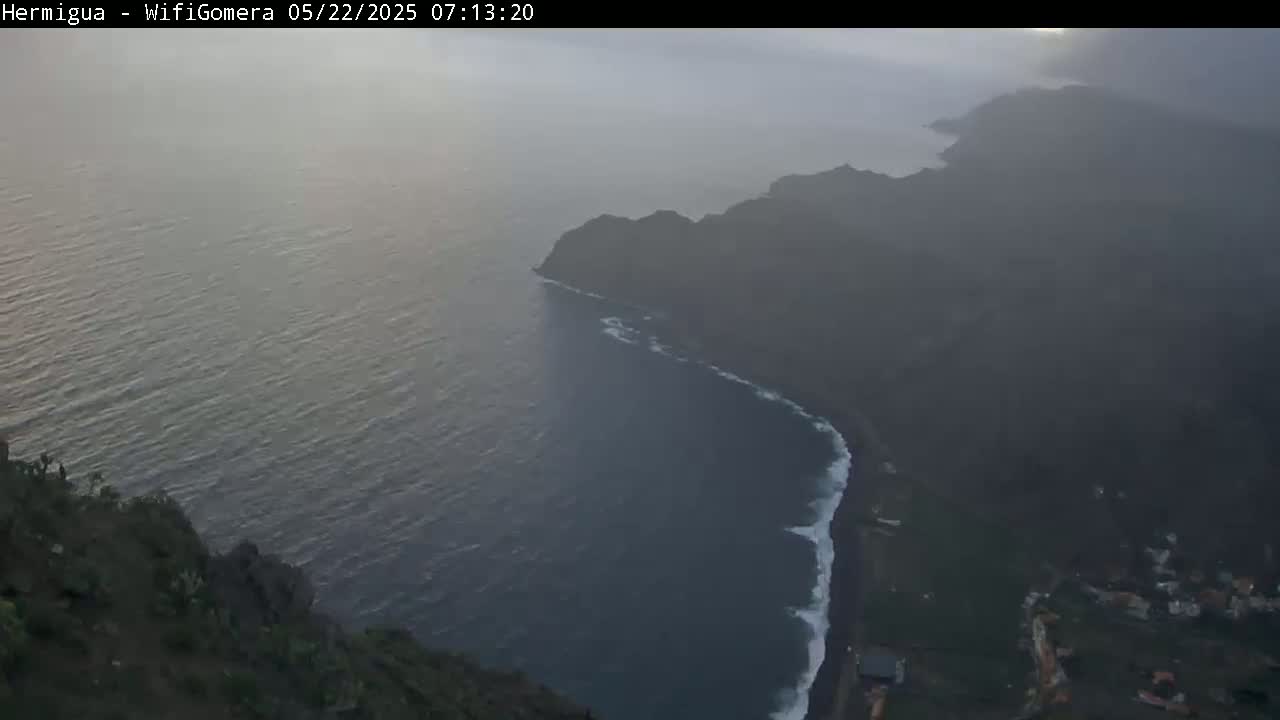 A hazy sunrise reveals a dark, rocky coastline meeting a dark-blue ocean with small waves.