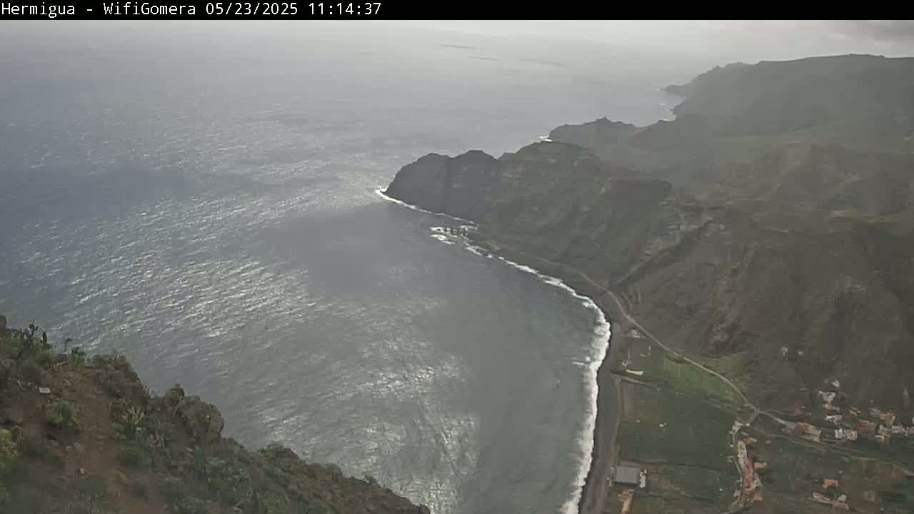 A high-angle view shows a dark-grey ocean meeting a rugged, dark-brown coastline under an overcast sky.
