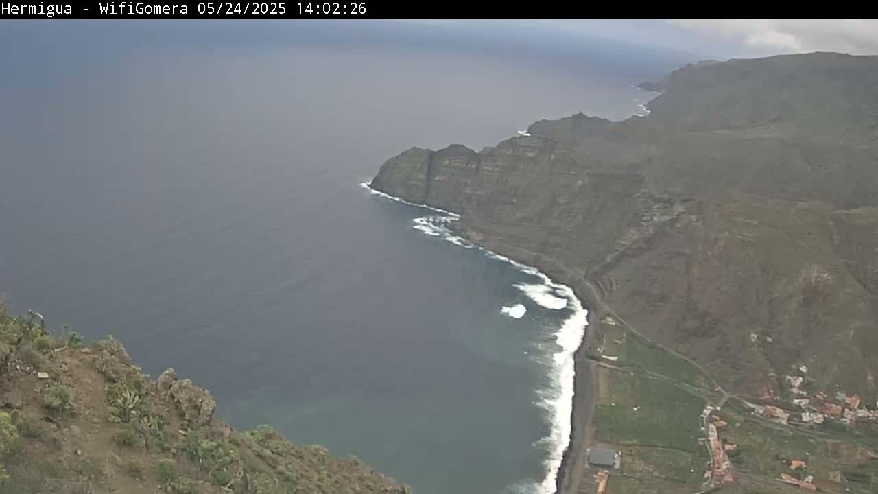 A high-angle view shows dark-grey, rocky cliffs meeting a dark-blue ocean with white waves along a black-sand beach under an overcast sky.