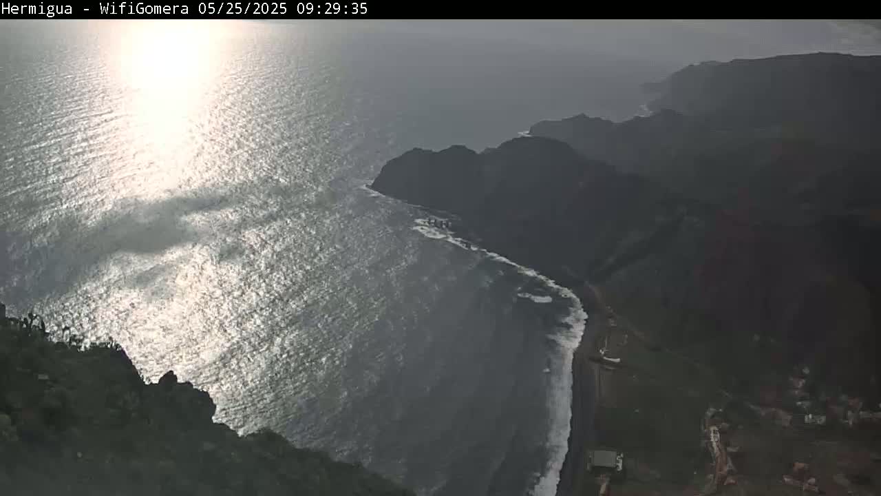 A high-angle, sunny view shows a dark rocky coastline meeting a sparkling ocean, with a small village visible at the bottom right.