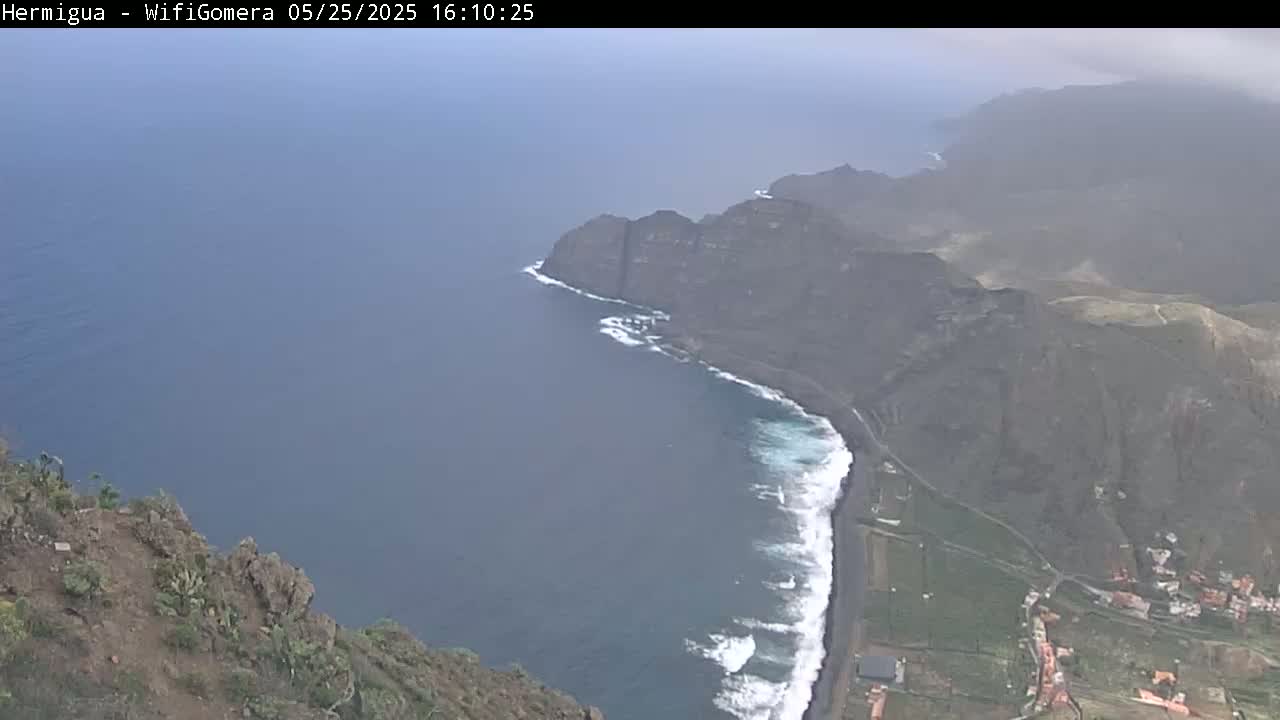 A hazy coastal view shows dark volcanic cliffs meeting a dark-blue ocean with white-capped waves, a black-sand beach, and a small village nestled at the cliff's base.