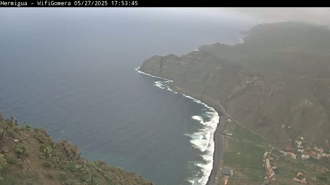 A high-angle view shows a hazy, overcast day overlooking a dark-sand beach meeting a dark-blue ocean with white-capped waves crashing on the shore, backed by a cliffside with some low-lying vegetation and buildings visible in the distance.