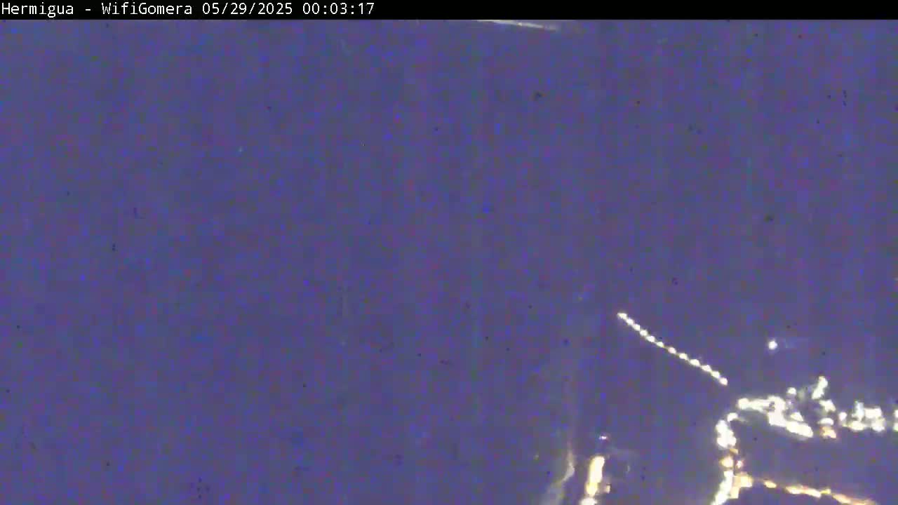 A dark, nighttime scene shows a distant cluster of lights at the bottom of the frame, possibly a town or city, with a faint streak of light crossing the upper portion of the image.