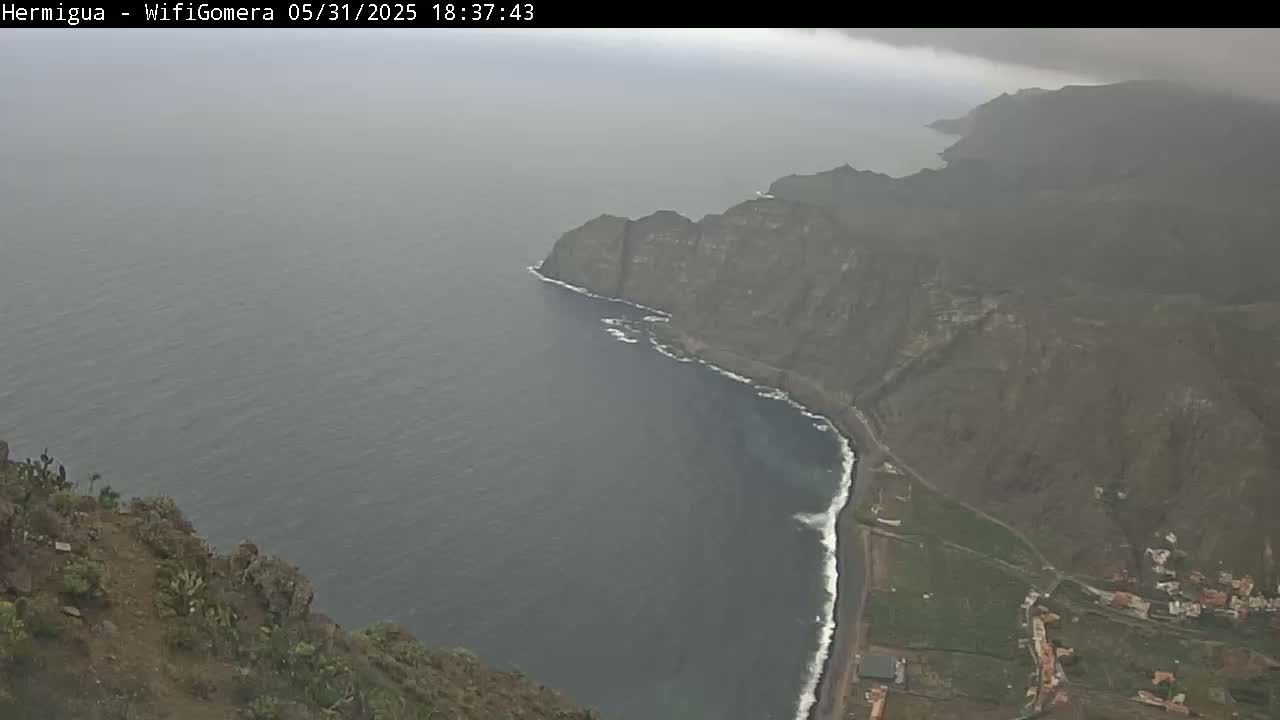 A coastal view shows dark, rocky cliffs meeting a hazy, gray ocean under an overcast sky.