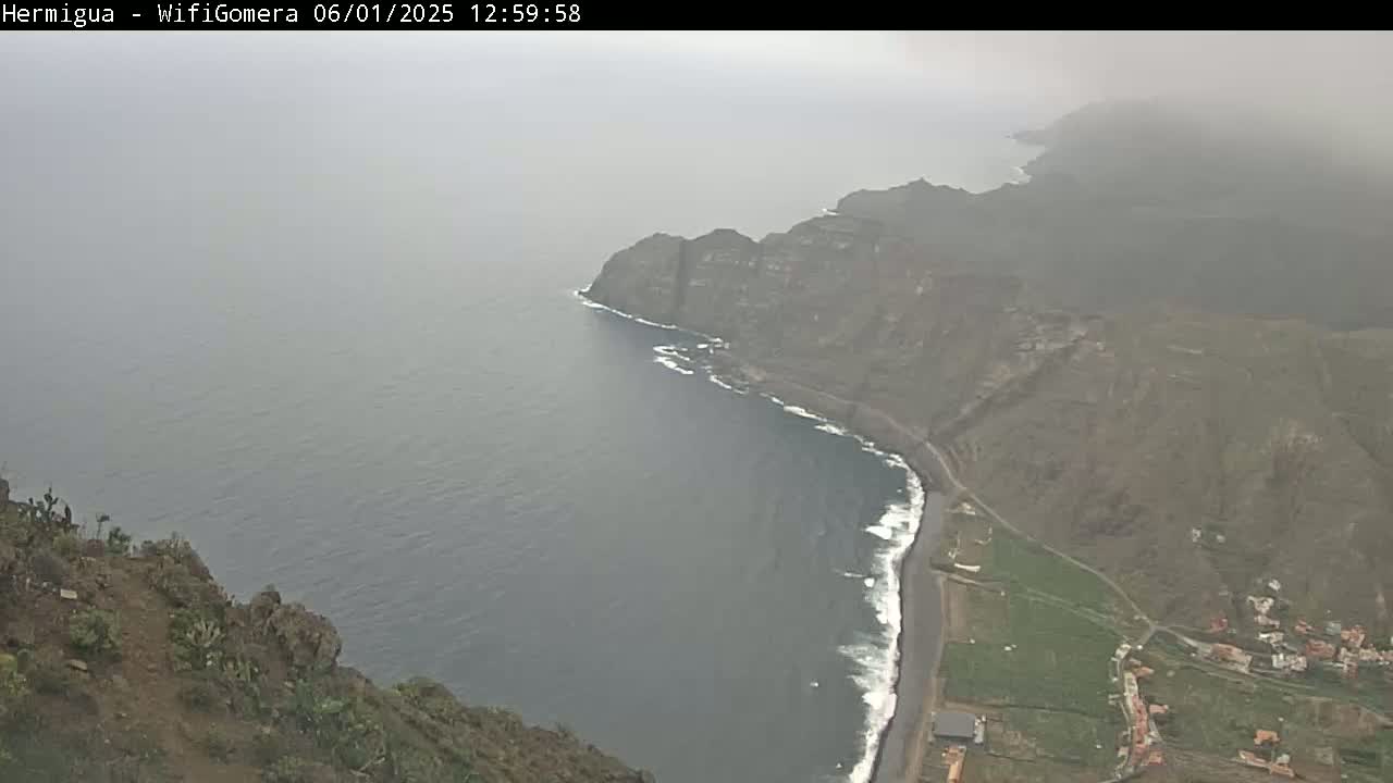 A hazy coastal view shows a dark-colored ocean meeting a rocky, brown cliffside with a small town and farmland visible, under an overcast sky.