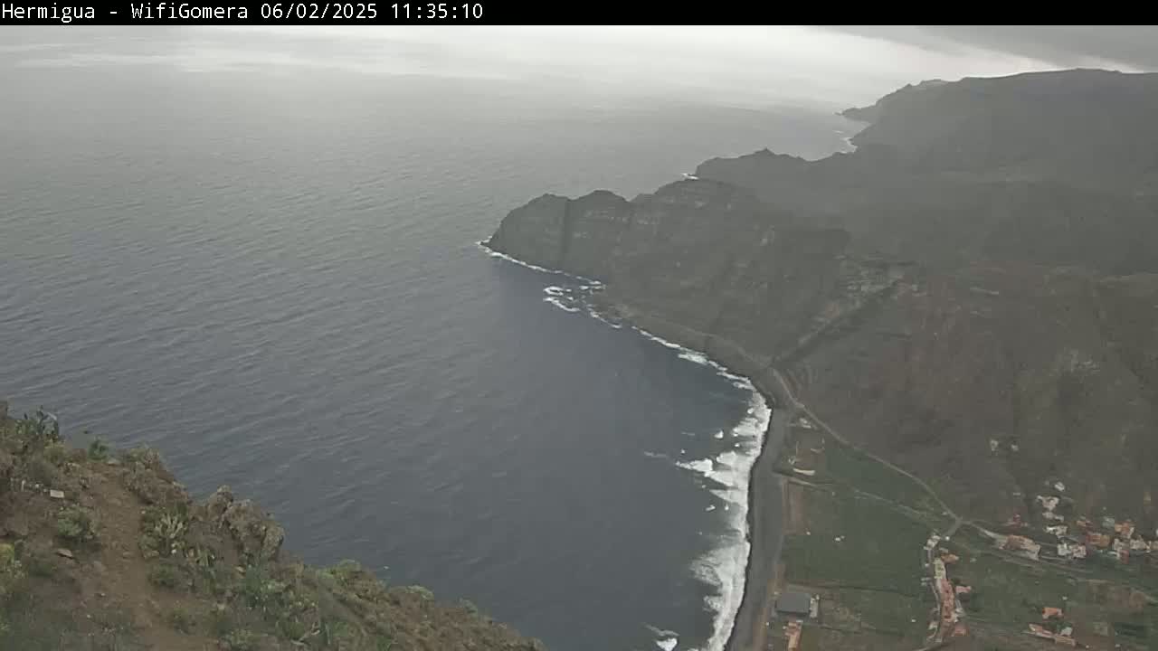 A high-angle, overcast view shows a dark-grey ocean meeting a dark-brown rocky coastline with a small amount of white-capped waves along the shore.