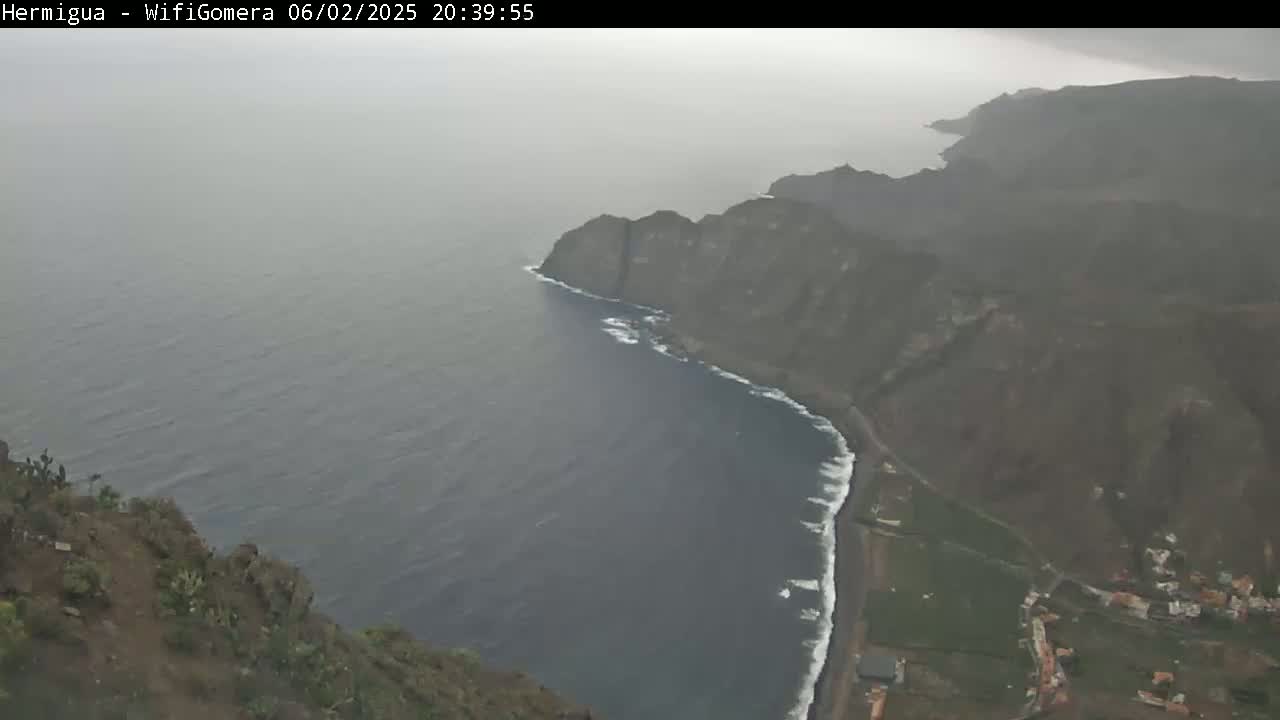 A hazy coastal view shows dark-colored cliffs meeting a dark-blue ocean with small waves, partially obscured by mist.