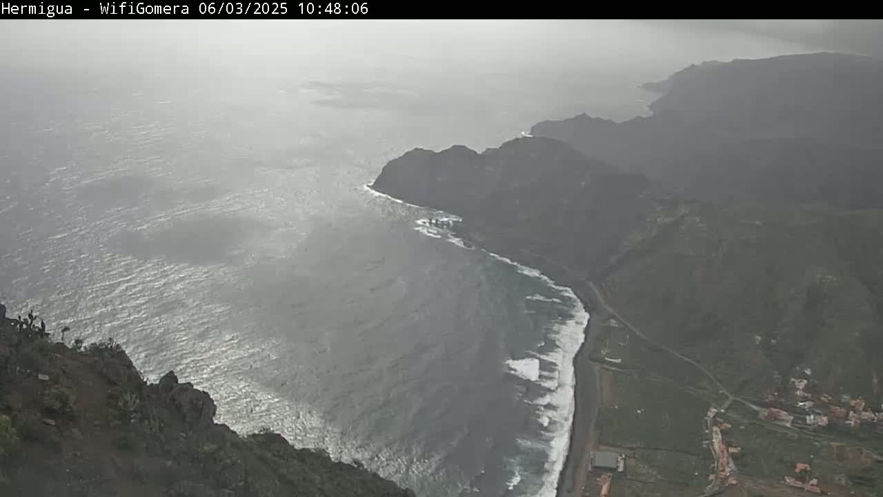 A high-angle view shows a rocky coastline with crashing waves on a hazy, overcast day.