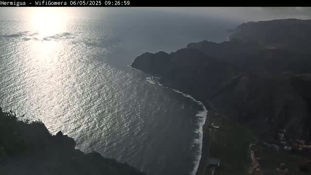 A high-angle view shows a sunlit ocean meeting dark, rocky cliffs and a shoreline with small waves under a partly cloudy sky.
