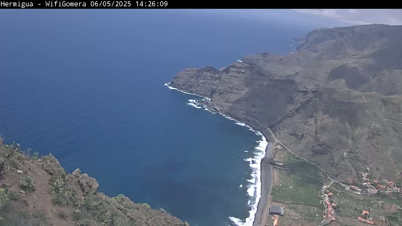 A high-angle view shows a dark-blue ocean meeting a rocky, brown coastline with a small beach and a village under a mostly clear sky.
