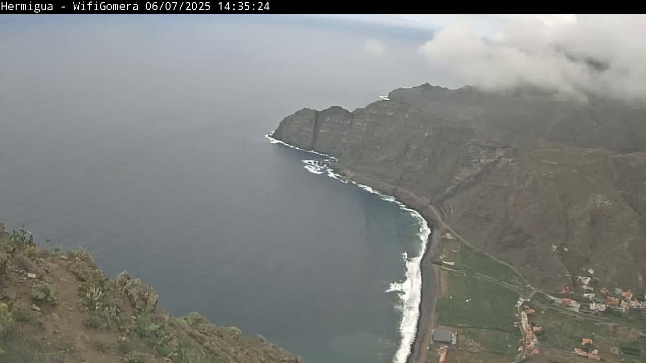 A high-angle view shows a dark-colored, sandy beach curving into a dark-blue ocean next to a steep, rocky cliff and low-lying land, all under a cloudy sky.