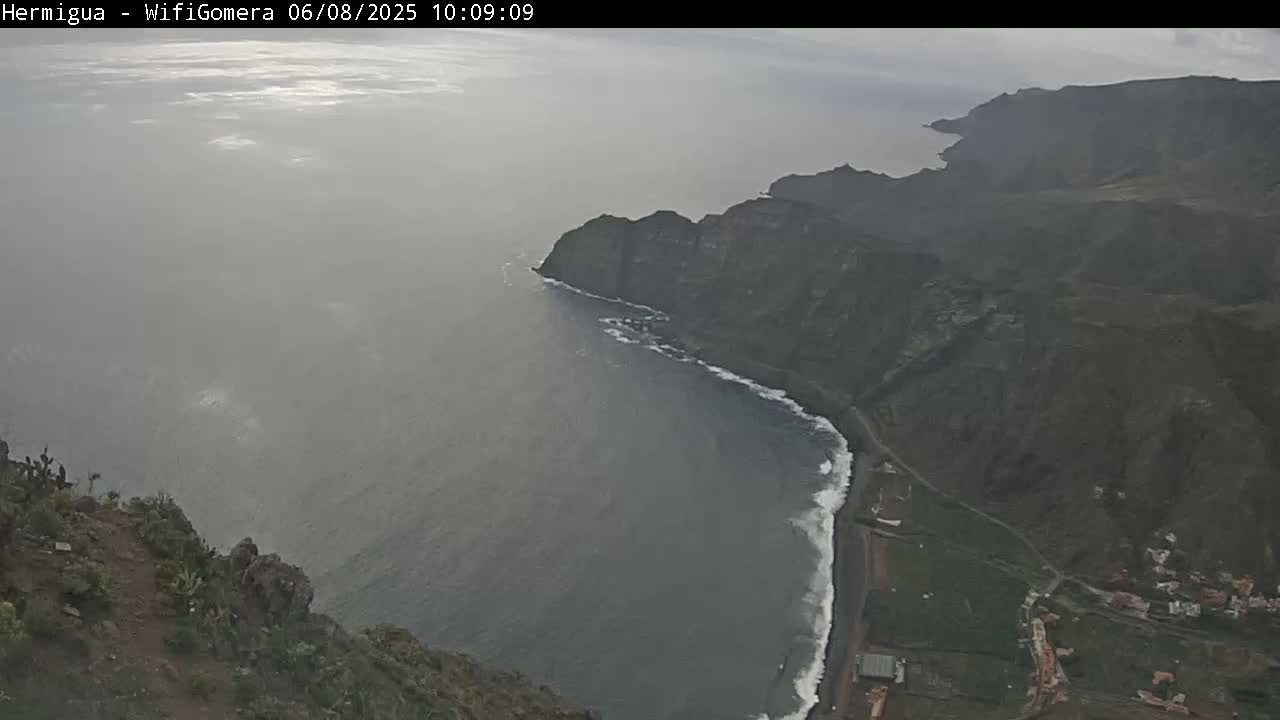 A high-angle view shows a coastline with dark cliffs, a gray ocean with small waves, and a hazy sky.