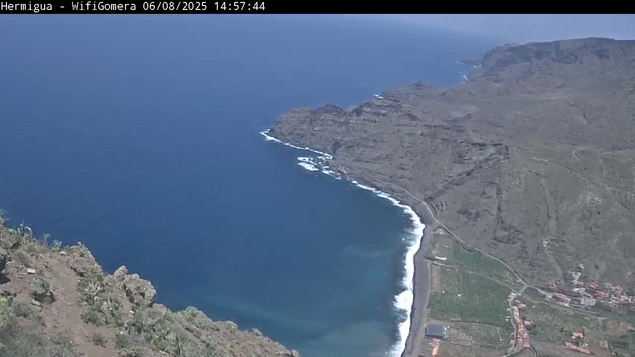 A high-angle view shows a dark-colored rocky coastline meeting a deep blue ocean under a clear sky.