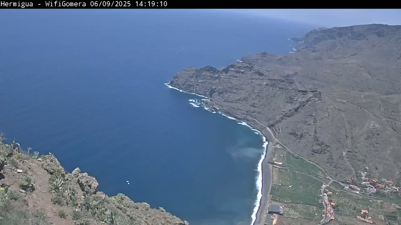 A high-angle view shows a dark-colored, rocky coastline meeting a deep blue ocean under a clear sky.