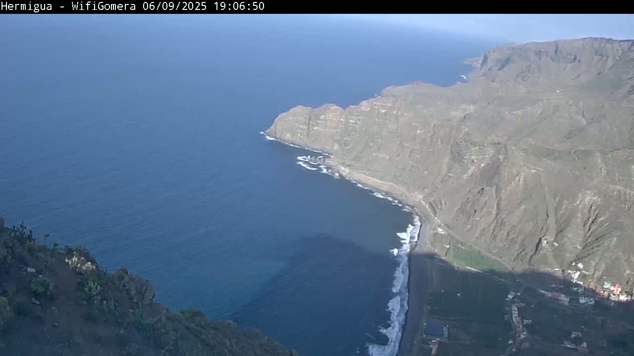 A high-angle view shows a dramatic coastline with dark cliffs meeting a calm, deep blue ocean under a clear sky.