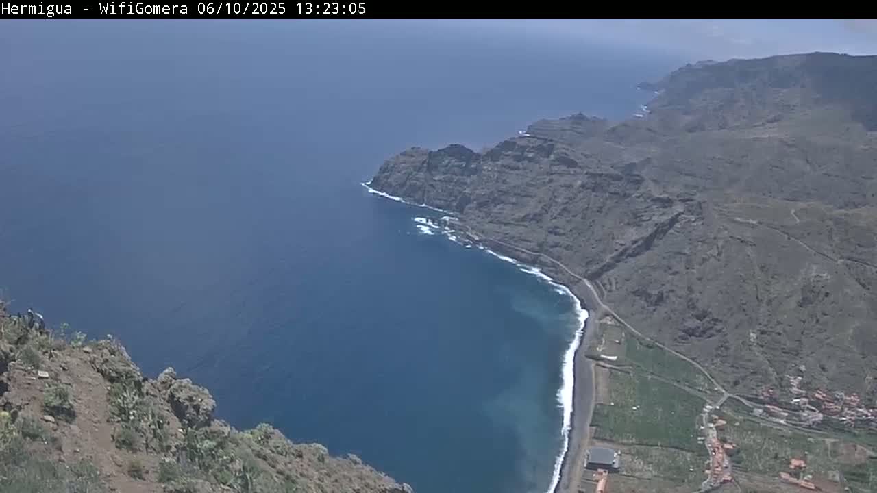 A high-angle view shows a dark-colored, rocky coastline meeting a deep blue ocean under a clear sky.
