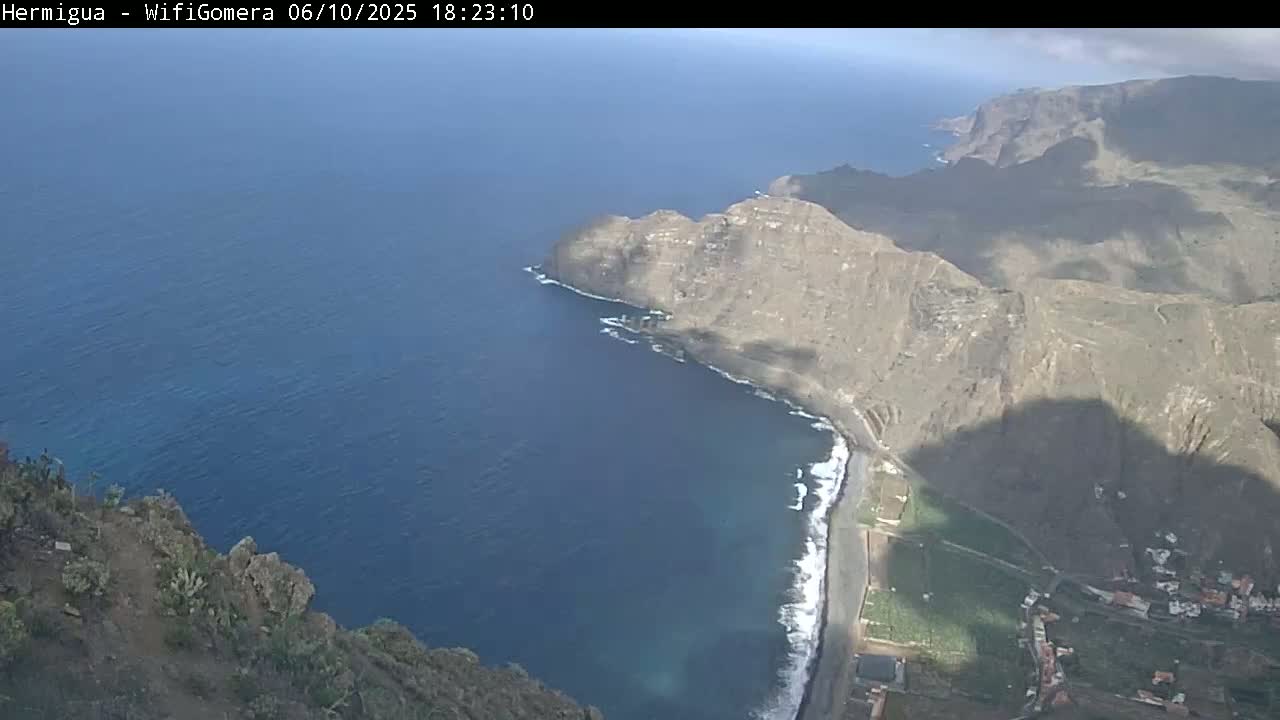 A high-angle view shows a rocky coastline with a small beach, dark blue ocean, and mountainous terrain under partly cloudy skies.