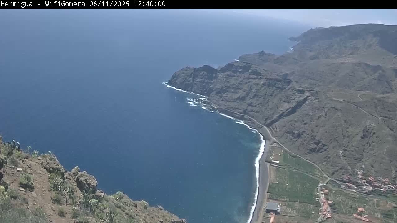 A high-angle view shows a dark-colored, rocky coastline meeting a deep blue ocean under a clear sky.