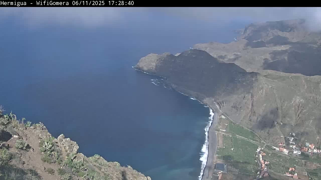A high-angle view shows a rugged, mountainous coastline meeting a dark blue ocean under a mostly clear sky.