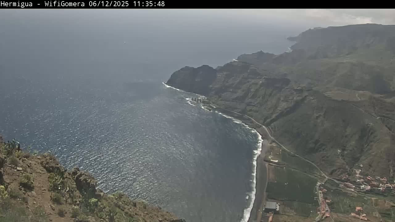 A high-angle view shows a rocky, dark-colored coastline meeting a somewhat choppy ocean under a hazy sky.