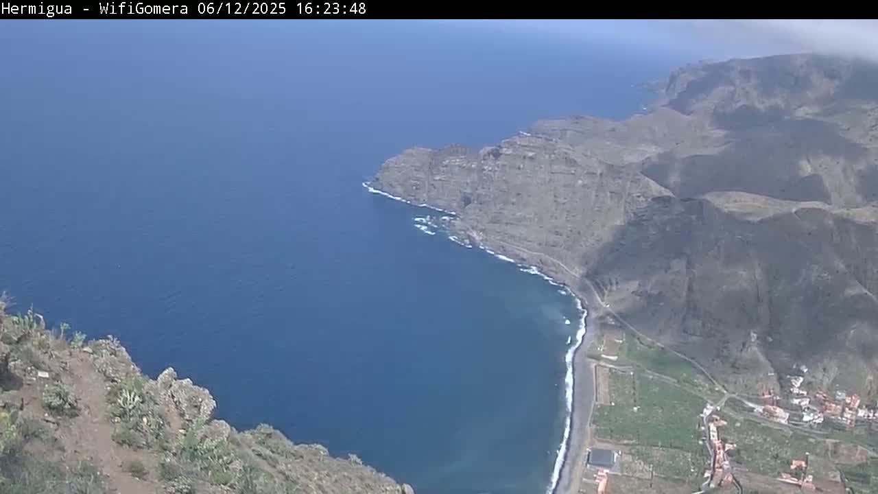 A high-angle view shows a dark-blue ocean meeting a rugged, brown coastline under a mostly clear sky.