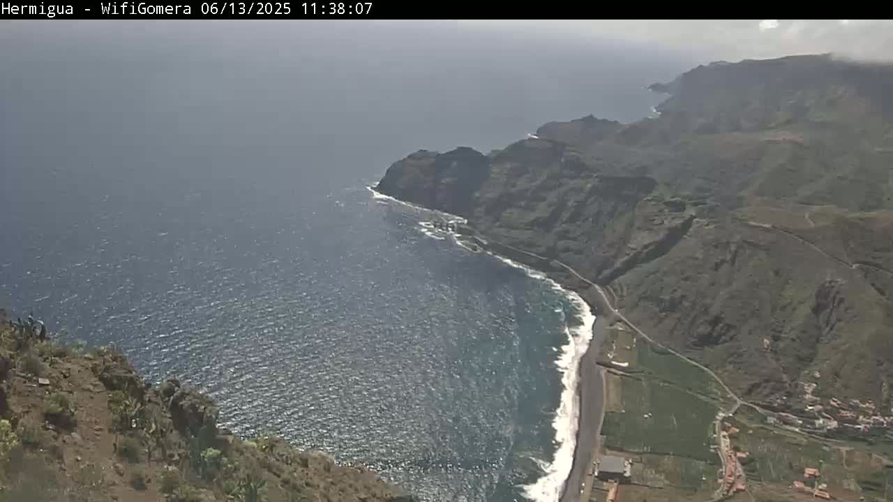 A high-angle view shows a rocky, dark-colored coastline meeting a wavy ocean under a mostly cloudy sky.
