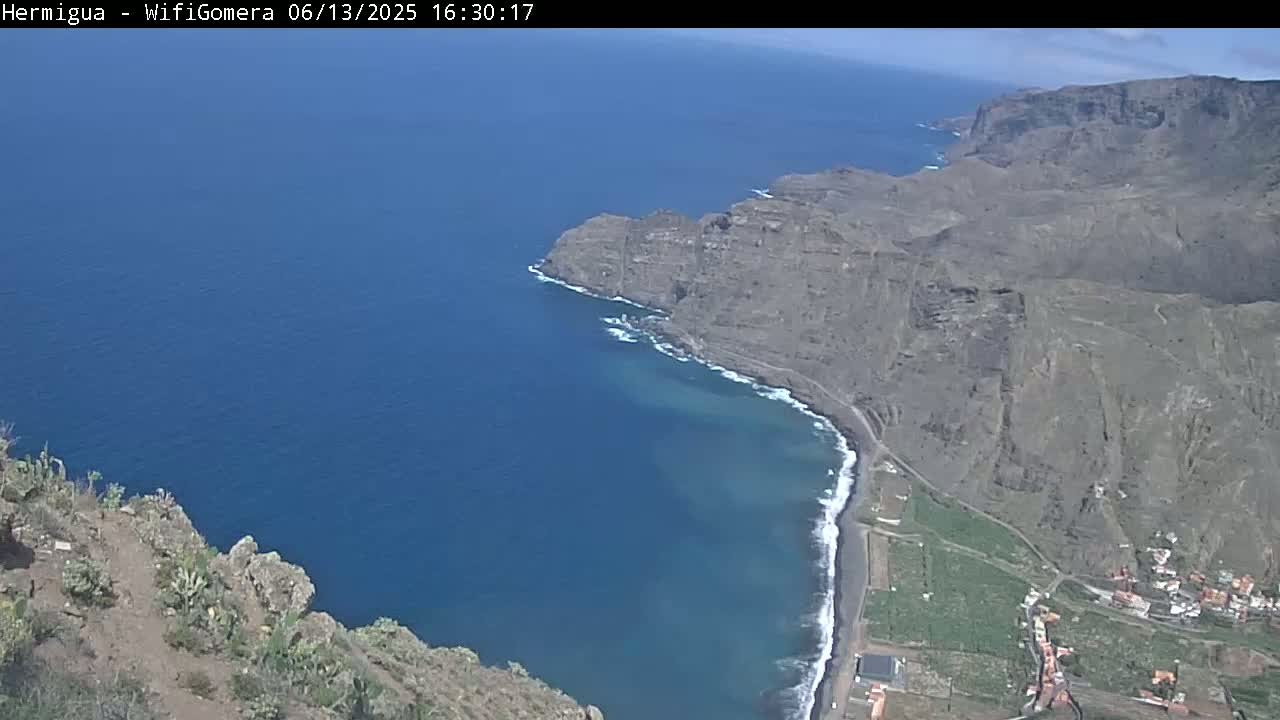A high-angle view shows a rocky, dark-colored coastline meeting a deep blue ocean under a mostly clear sky.