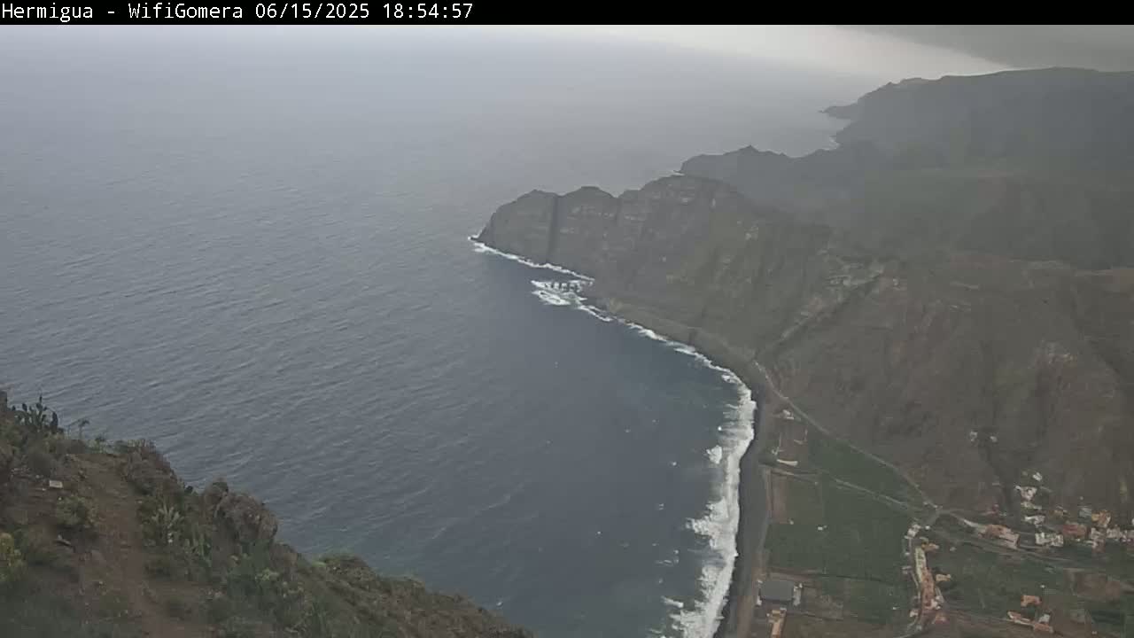 A high-angle view shows a dark, rocky coastline meeting a hazy, gray ocean under an overcast sky.