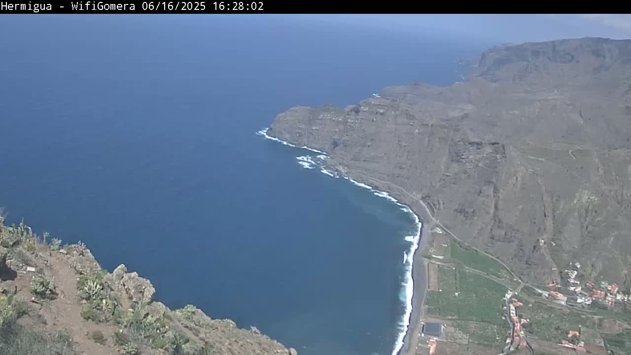 A high-angle view shows a rocky coastline with a dark-colored beach curving into a deep blue ocean under a clear, sunny sky.