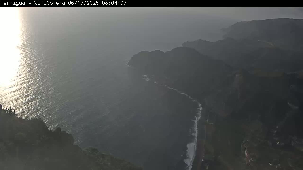 A hazy, sunlit ocean meets dark, mountainous cliffs under a mostly cloudy sky.