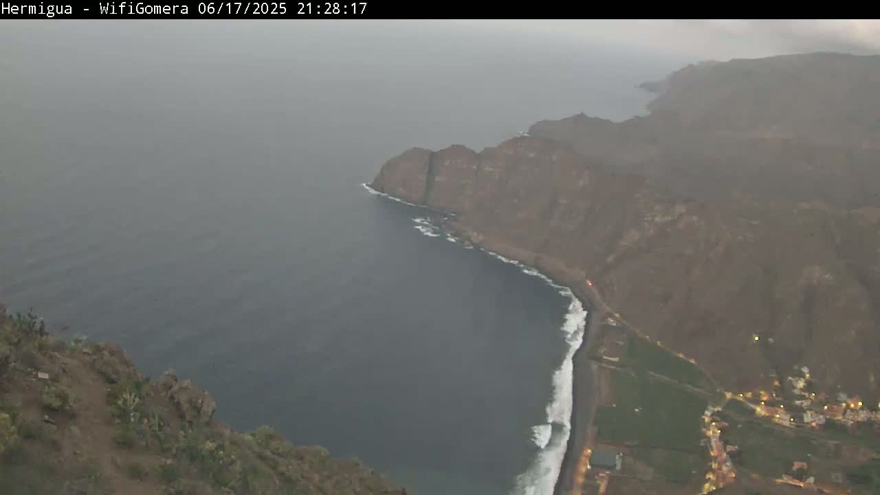 A hazy, high-angle view shows a dark-colored ocean meeting a rocky, mountainous coastline with a few lights visible at the base of the cliffs.