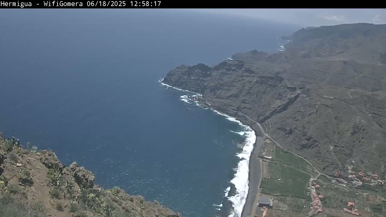 A high-angle view shows a dark-colored, rocky coastline with a dark-sand beach meeting a deep blue ocean under a bright, sunny sky.