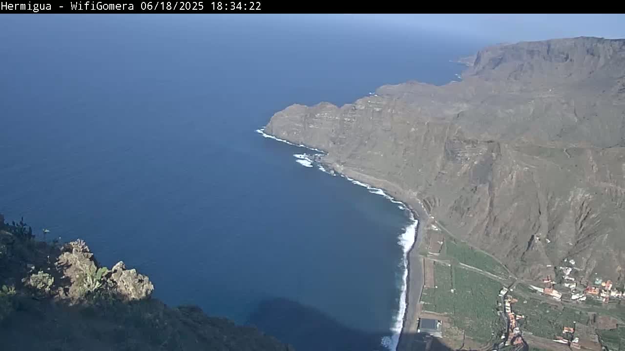 A high-angle, long shot shows a dark-blue ocean meeting a rocky, brown coastline with a small town nestled at the base of steep cliffs under a mostly clear sky.