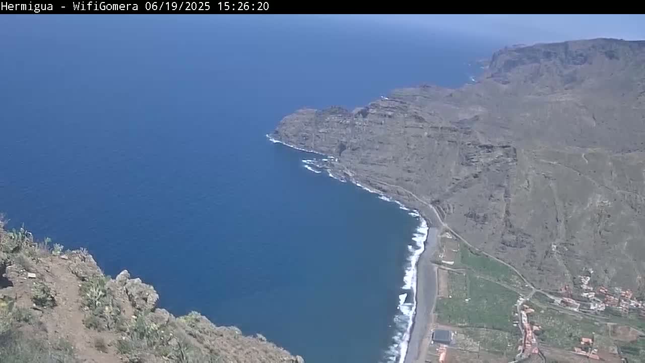 A high-angle view shows a rocky, arid coastline meeting a deep blue ocean under a clear sky.