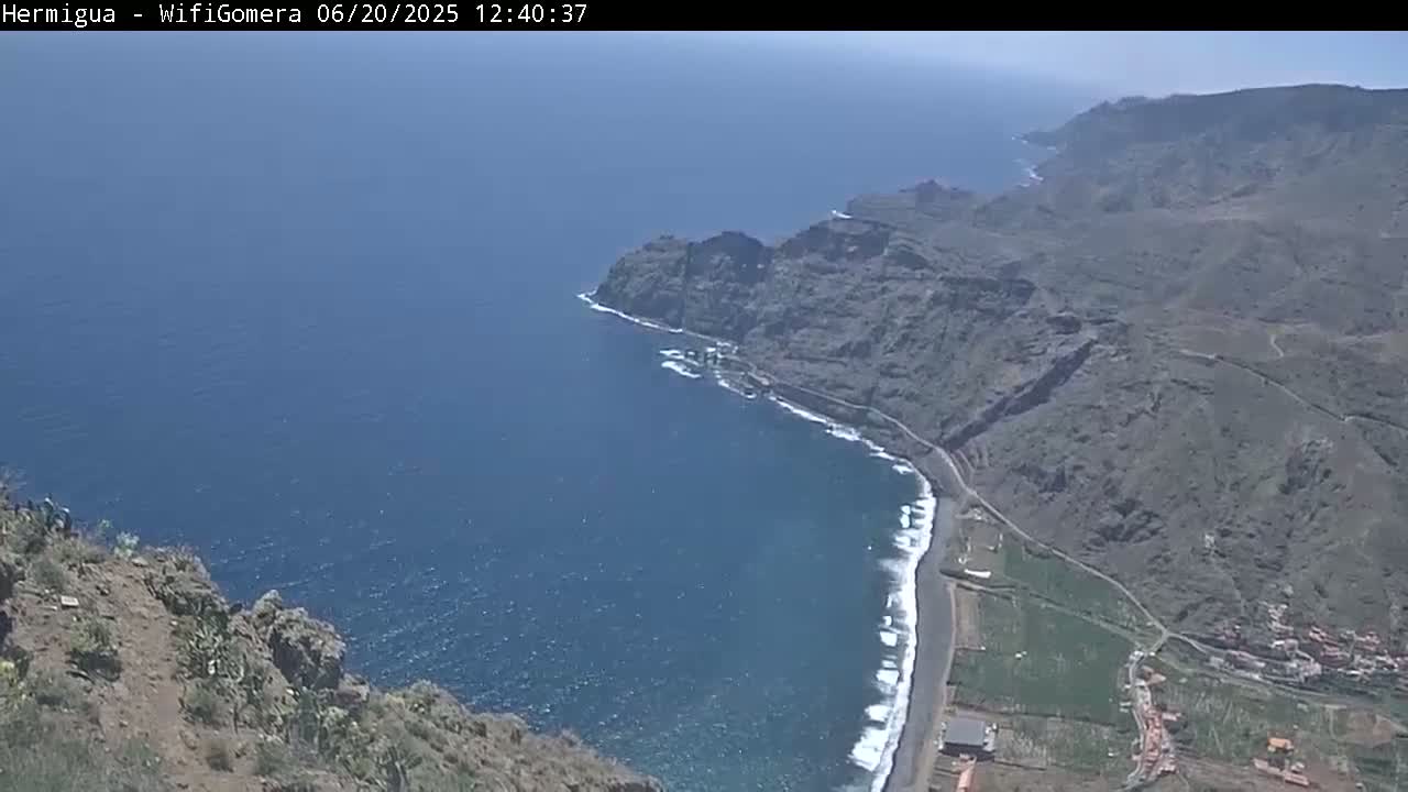 A high-angle view shows a rocky, mountainous coastline meeting a dark blue ocean on a sunny day.