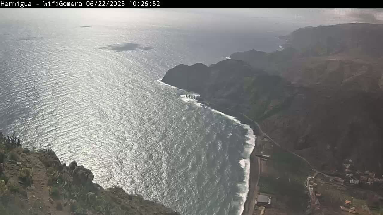 A high-angle view shows a rocky coastline meeting a somewhat wavy ocean under hazy skies.