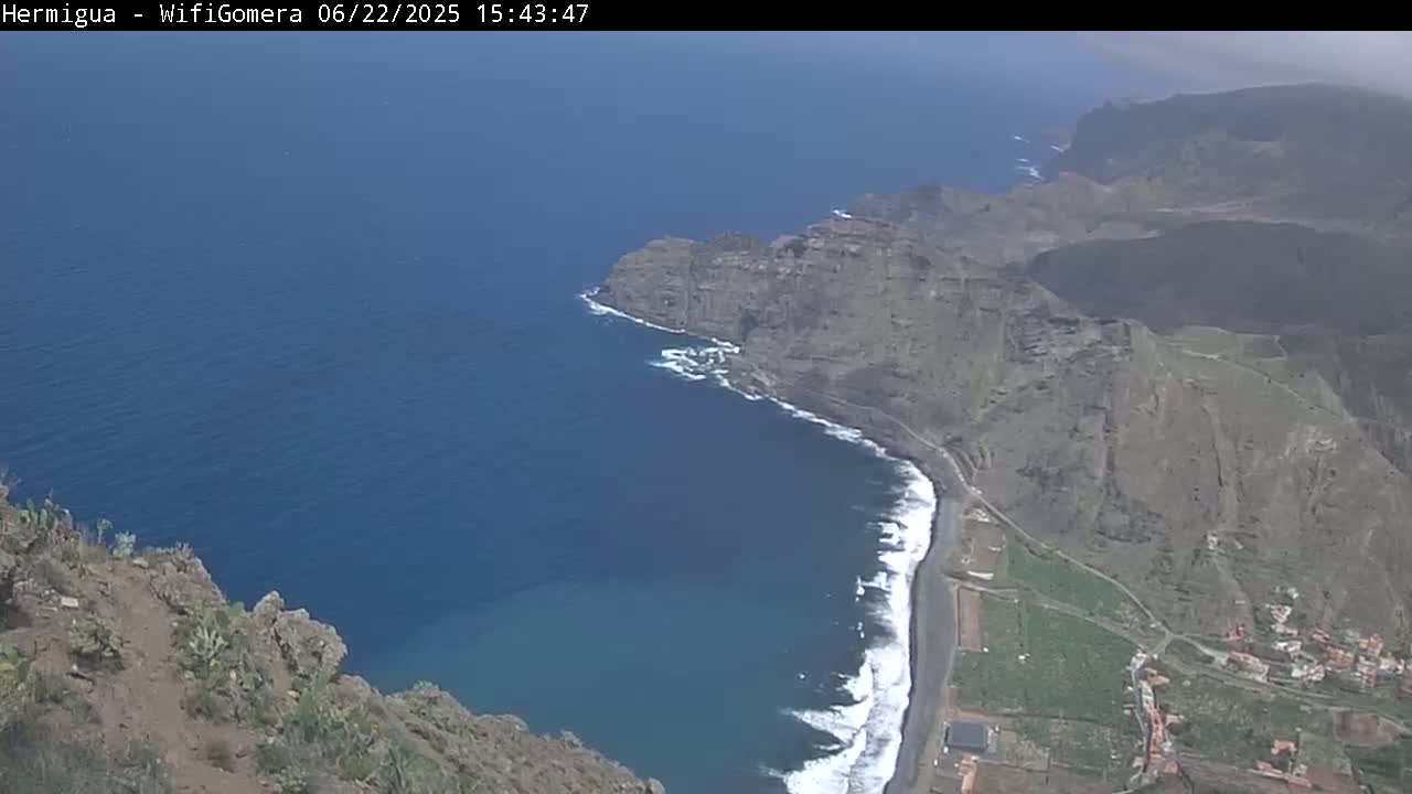 A high-angle view shows a dark-colored beach curving into a deep blue ocean next to a rocky, brown cliffside under a mostly clear sky.