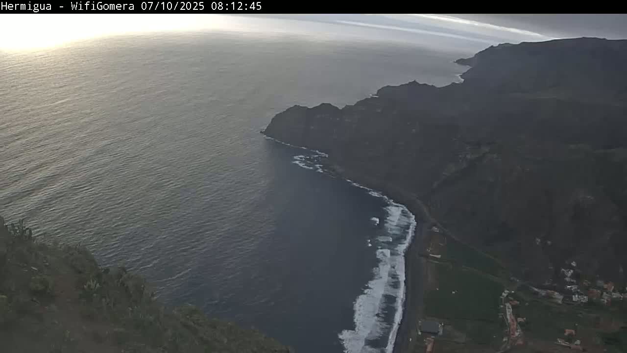 An aerial view shows a dark, rocky coastline with waves breaking on a dark-sand beach, under an overcast sky.