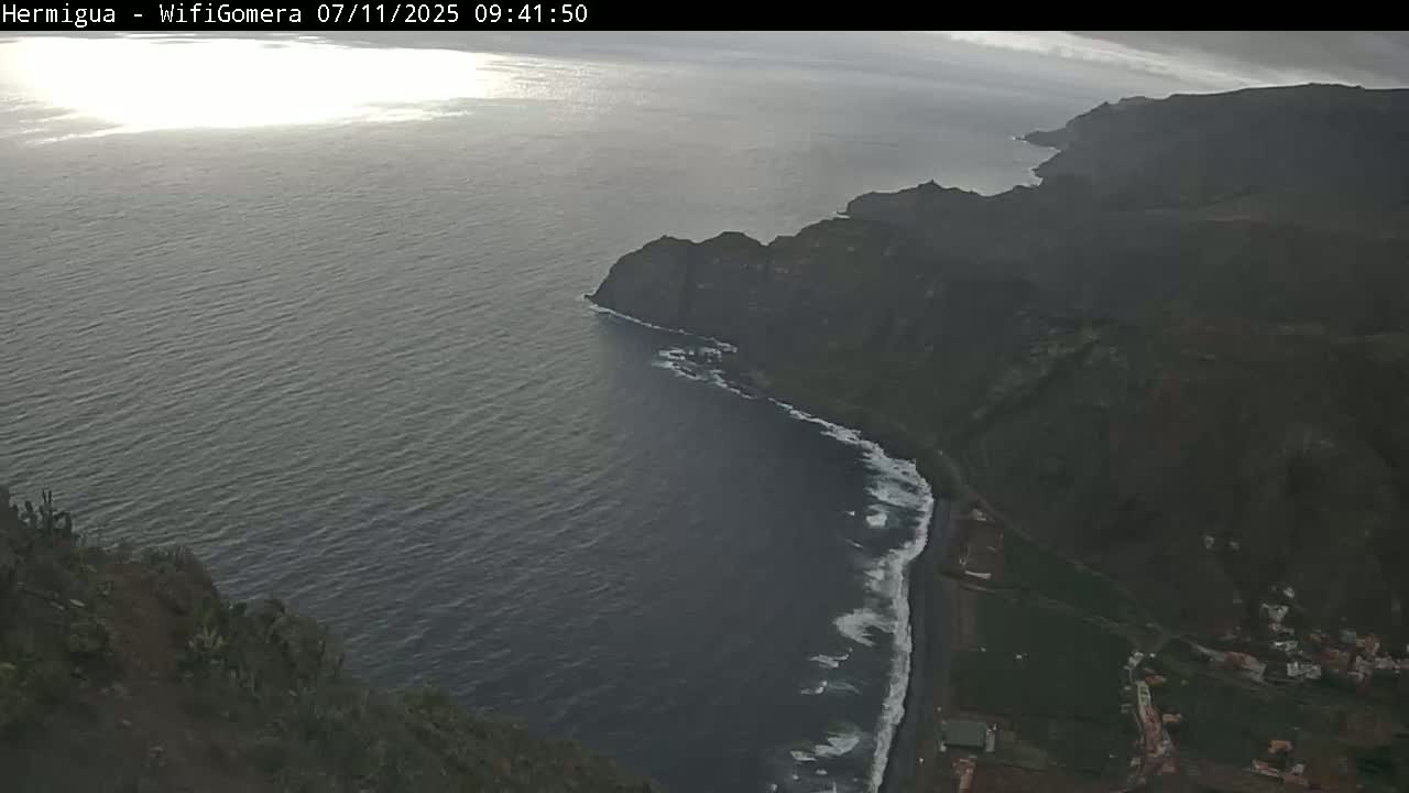 A dark, overcast sky looms over a dramatic coastline where dark-colored cliffs meet a dark ocean with white-capped waves along a black-sand beach.